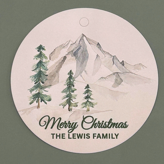 A round hanging gift tag with a watercolor mountain and pine tree design, featuring the text 'Merry Christmas and personalized name