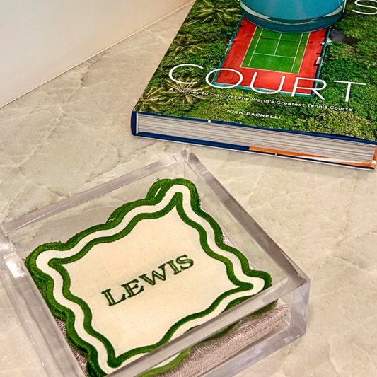 A personalized clear acrylic square tray holding a cream-colored fabric linen napkin with green trim and the name in green lettering, styled on a light marble surface.