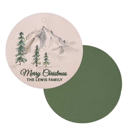 A round Christmas tag featuring a watercolor mountain and pine tree illustration above the text 'Merry Christmas personalized with family name
