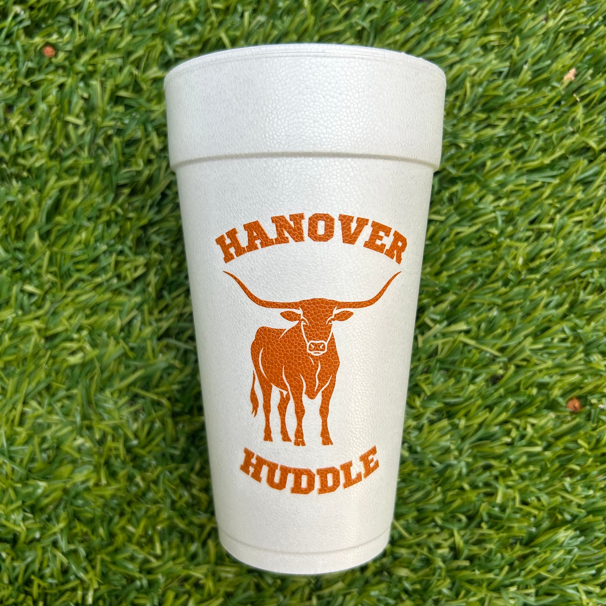 A white 20 ounce foam cup with a custom orange graphic of a longhorn steer and the text  printed on it, placed on green  turf.