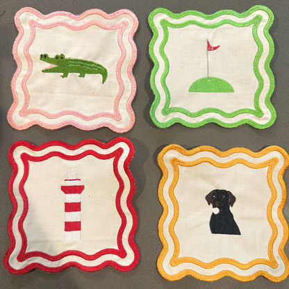 Four embroidered fabric napkins. Each coaster has a scalloped border and a different embroidered animal or object: a pink border with a green alligator, a green border with a golf hole and red flag, a red border with a red-and-white lighthouse, and a yellow-orange border with a black dog.