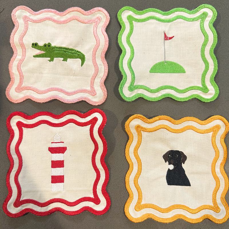 Four embroidered fabric napkins. Each coaster has a scalloped border and a different embroidered animal or object: a pink border with a green alligator, a green border with a golf hole and red flag, a red border with a red-and-white lighthouse, and a yellow-orange border with a black dog.