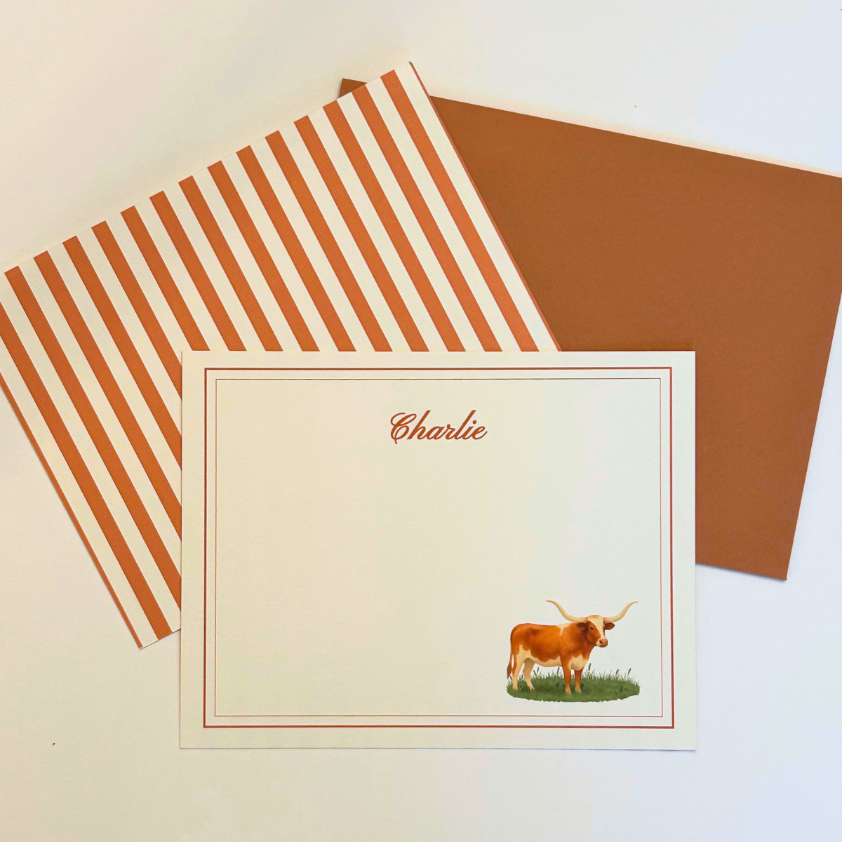 A personalized stationery set featuring a cream note card with a thin border, personalized name in script, and an illustration of a longhorn cow, shown with a orange envelope and a striped back