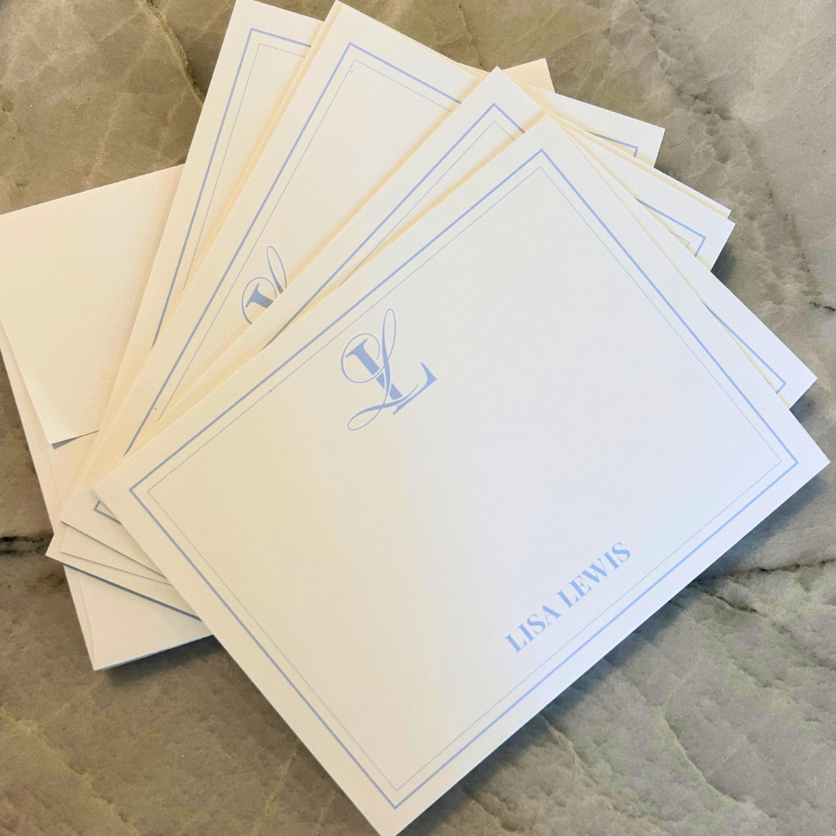 A stack of personalized stationery cards The cards are off-white with a thin blue border, a small blue monogram logo, and the name printed in blue 