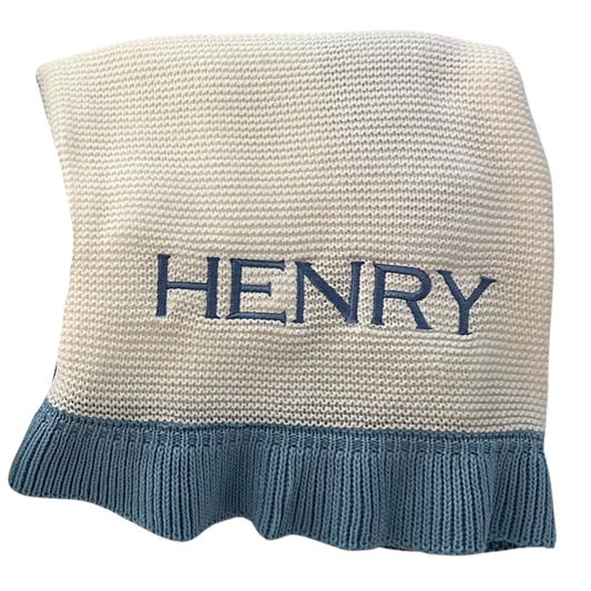 A personalized cream-colored knit baby blanket with a blue ruffled hem and the name embroidered in blue across the front.