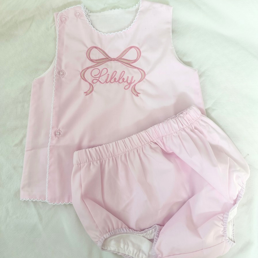 A baby girl's two-piece diaper set consisting of a light pink sleeveless top with trim, buttons and pink embroidered text  under a bow, paired with matching light pink bloomers