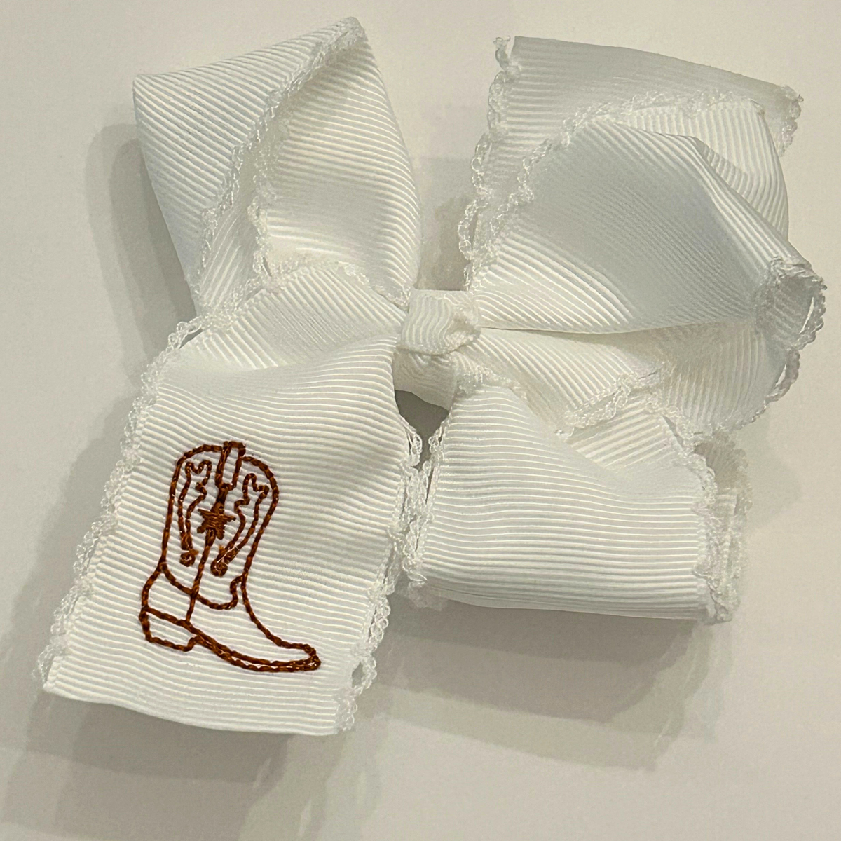 A white grosgrain ribbon hair bow with a western boot embroidered finished with a ruffled lace edge.