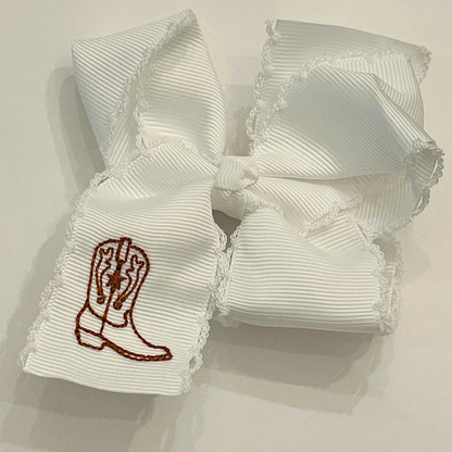 A white grosgrain ribbon hair bow with a western boot embroidered finished with a ruffled lace edge.