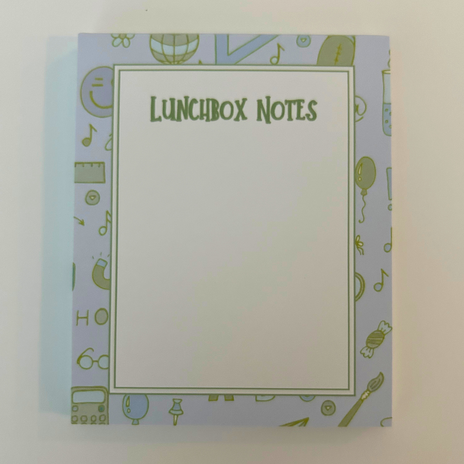 A notepad titled 'LUNCHBOX NOTES' with a large blank white center and a light blue border decorated with small green school-themed icons