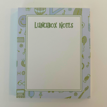A notepad titled 'LUNCHBOX NOTES' with a large blank white center and a light blue border decorated with small green school-themed icons